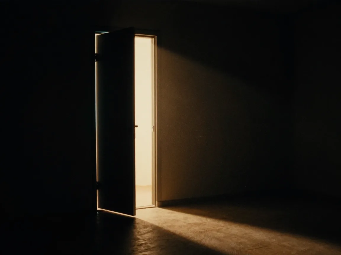 A narrow door standing open in a dark room, warm light falling through it onto the floor.